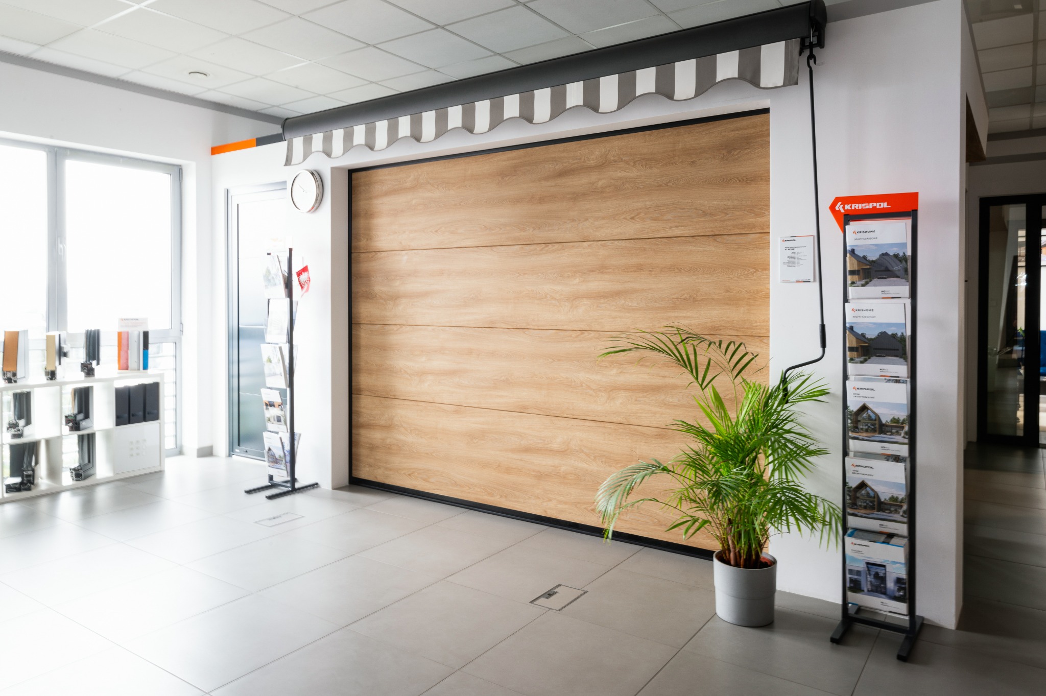 Jasna brama garażowa z drewnianym wzorem, z szarą markizą w paski. Obok roślina doniczkowa i stojak z ulotkami firmy Krispol. Wnętrze salonu ekspozycyjnego.