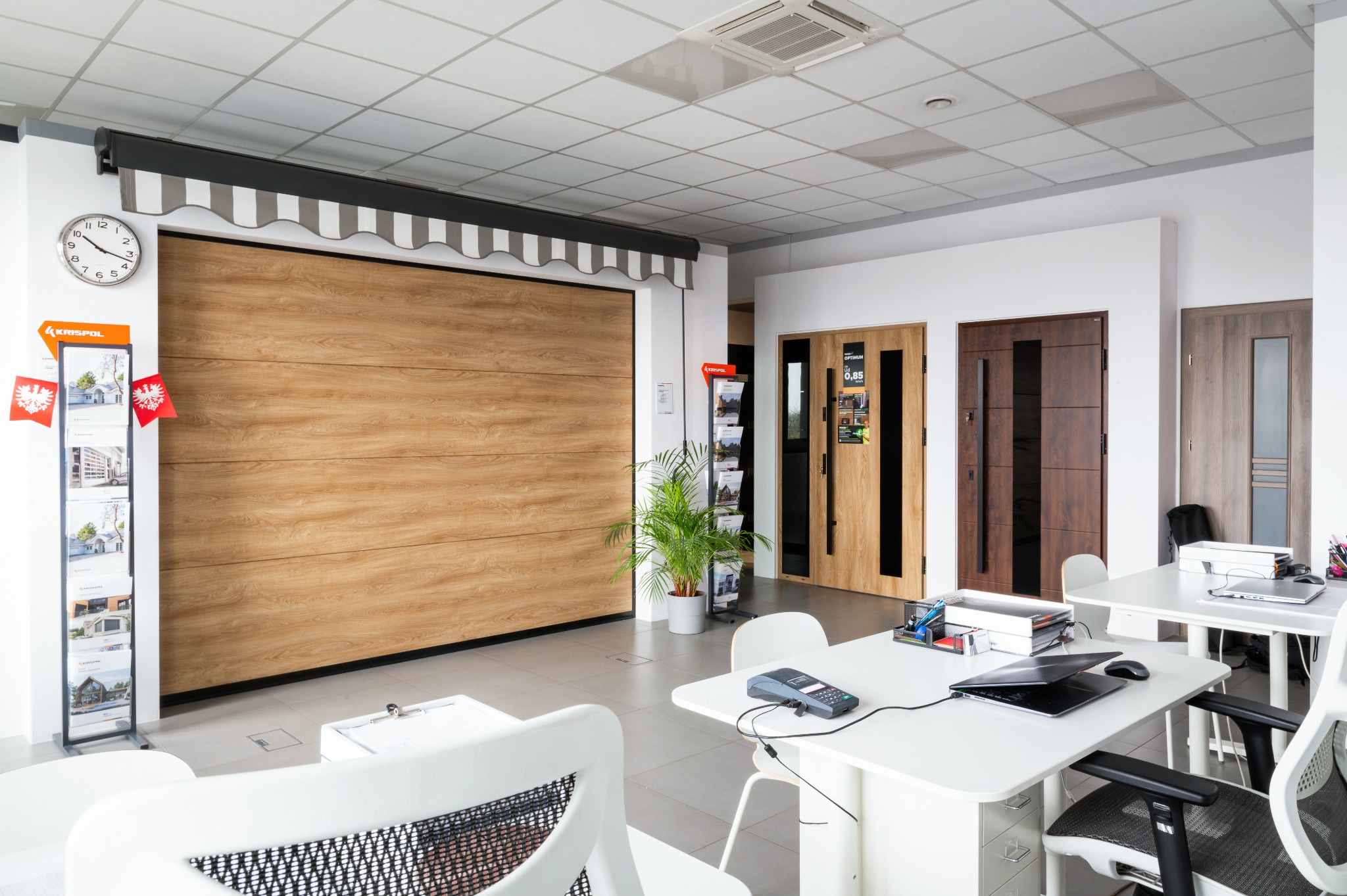 Ekspozycja: drewnopodobna brama garażowa z markizą w salonie, obok drzwi wewnętrzne i materiały reklamowe Krispol. Jasne wnętrze, nowoczesne meble.