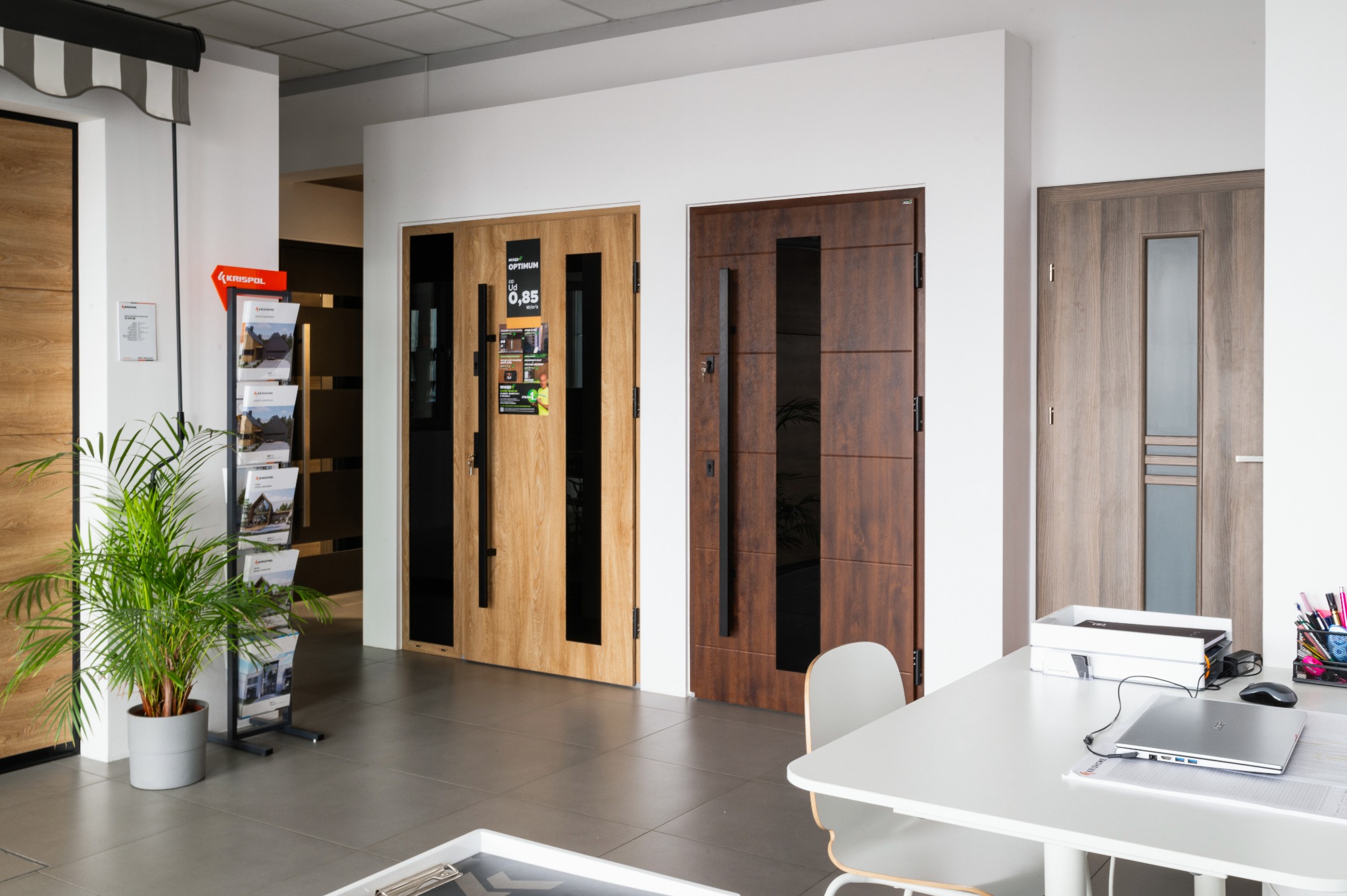 Ekspozycja trzech różnych modeli drzwi zewnętrznych w salonie, z widocznymi detalami wykończenia i okuć, uzupełniona o roślinę doniczkową i materiały reklamowe.