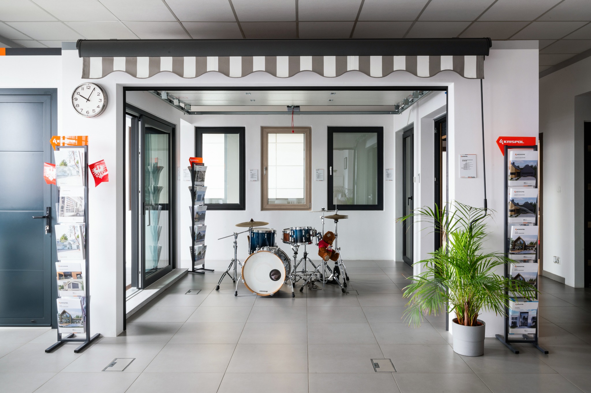 Ekspozycja salonowa: markiza w szaro-białe pasy nad oknami, drzwi wejściowe, stojaki z ulotkami i zegar na ścianie, uzupełniona o zestaw perkusyjny i roślinę doniczkową.