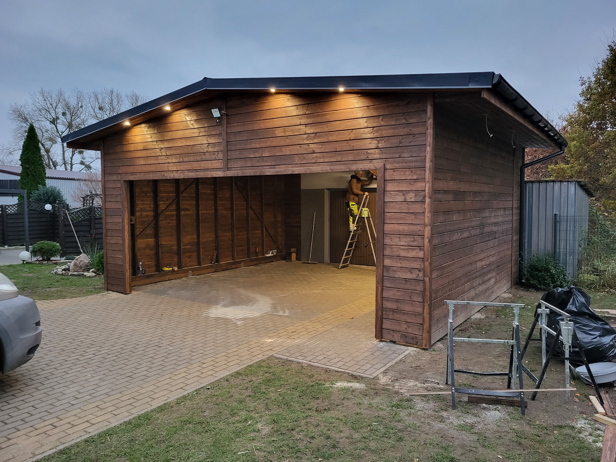 Drewniana konstrukcja garażu z oświetleniem zewnętrznym, pracownik na drabinie wewnątrz, widoczne narzędzia i materiały budowlane na zewnątrz, kostka brukowa przed garażem.