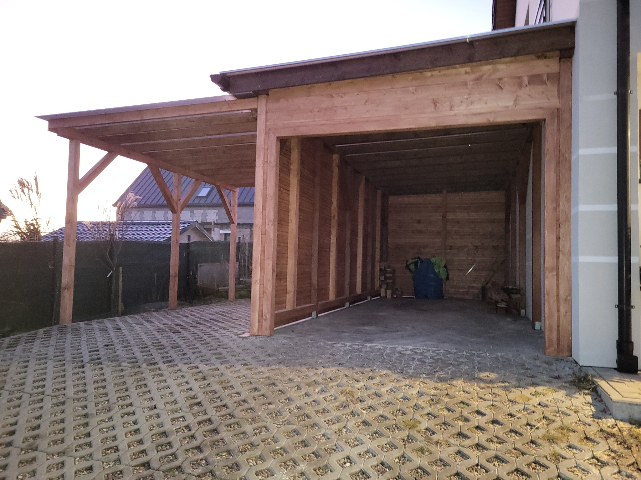 Drewniana wiata garażowa z podjazdem z ażurowych płyt betonowych, widok na sąsiedni dom z szarą dachówką.