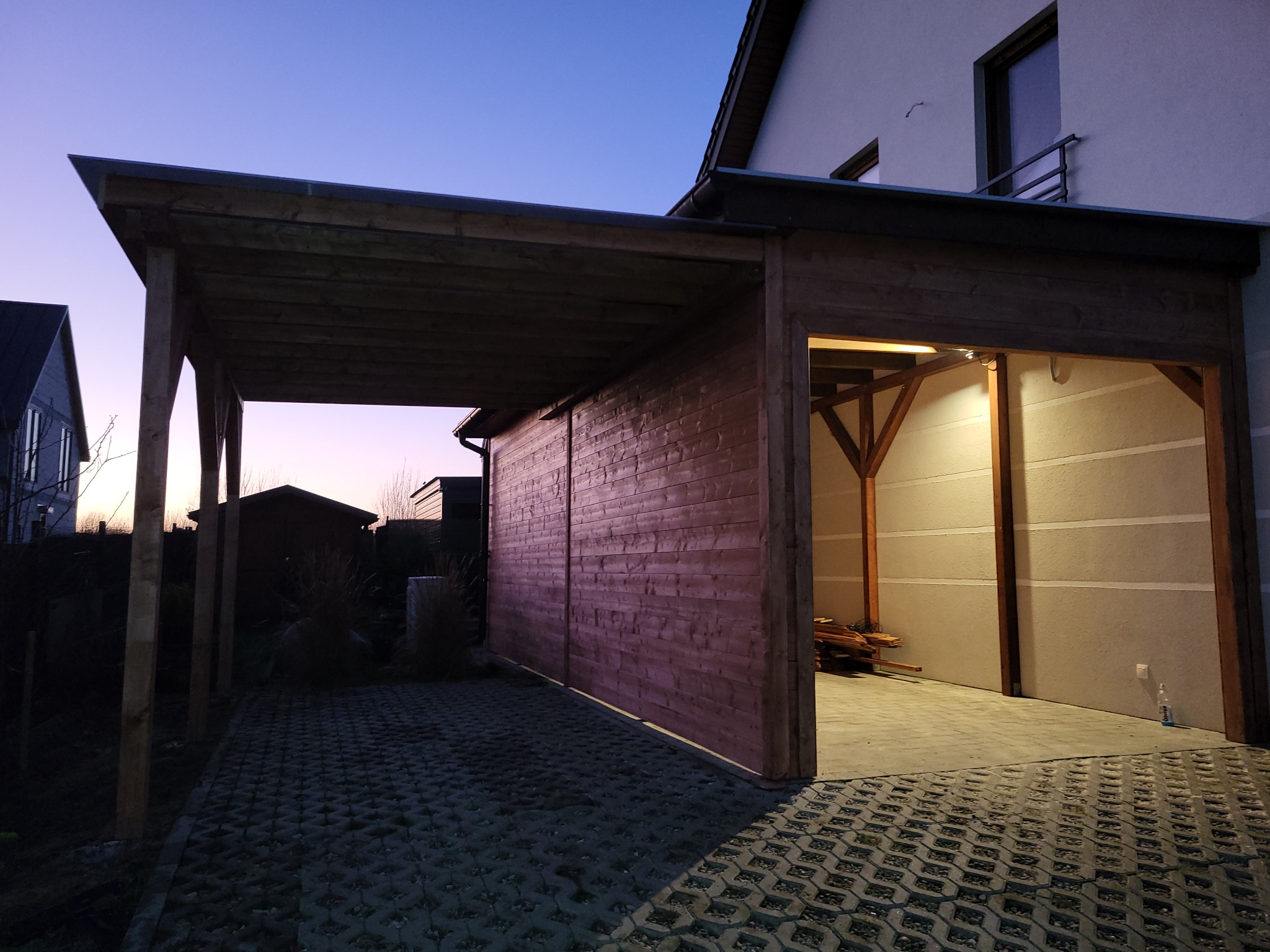 Zadaszone miejsce parkingowe z drewna, wykończone deskami, z otwartym wjazdem i podjazdem z ażurowych płyt betonowych, sfotografowane o zmierzchu.