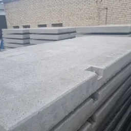 Stos szarych płyt drogowych z wycięciami na krawędziach, ułożonych na zewnątrz, z budynkiem z cegły w tle.