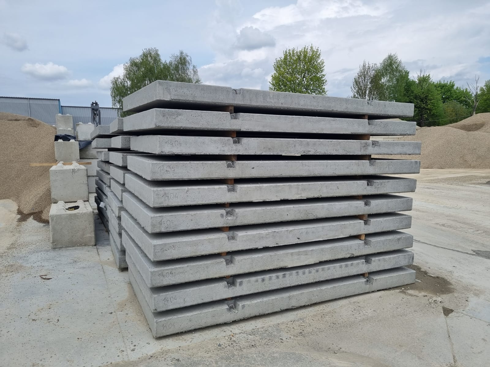Stos szarych, betonowych płyt drogowych z numerem '1404' ułożonych na drewnianych legarach, w tle hałda żwiru i fragment budynku.