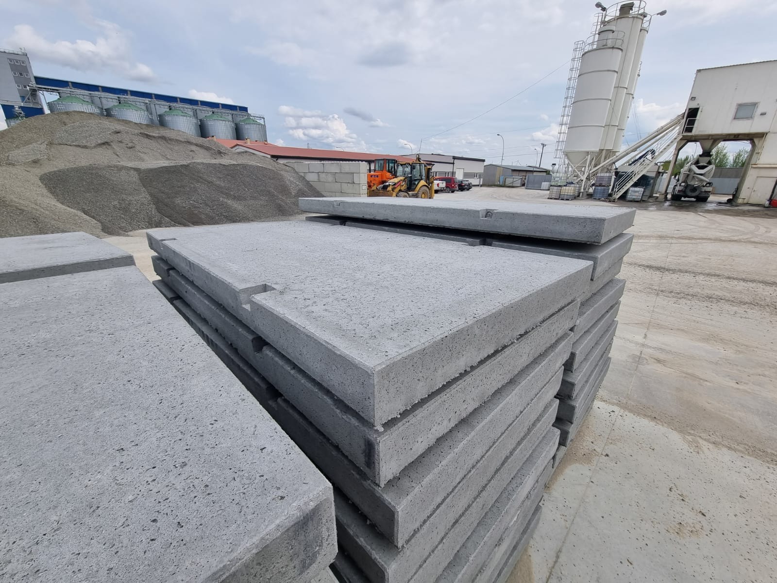 Stos szarych prefabrykowanych płyt betonowych z wycięciami, na tle plac budowy z koparką i silosami na kruszywo.