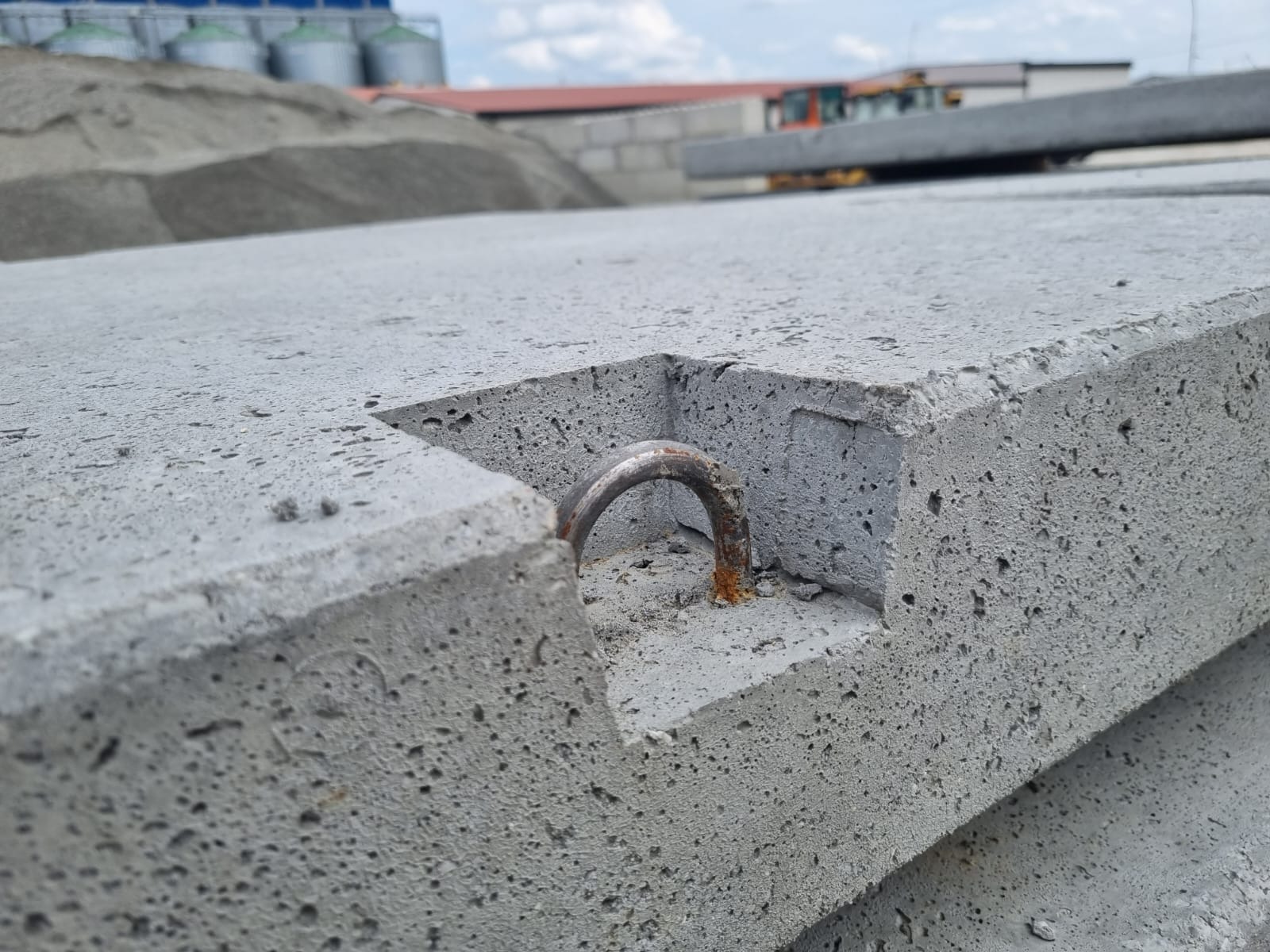 Szare płyty betonowe ułożone jedna na drugiej, widoczny uchwyt montażowy z lekką rdzą, w tle hałda piasku i budynki przemysłowe.