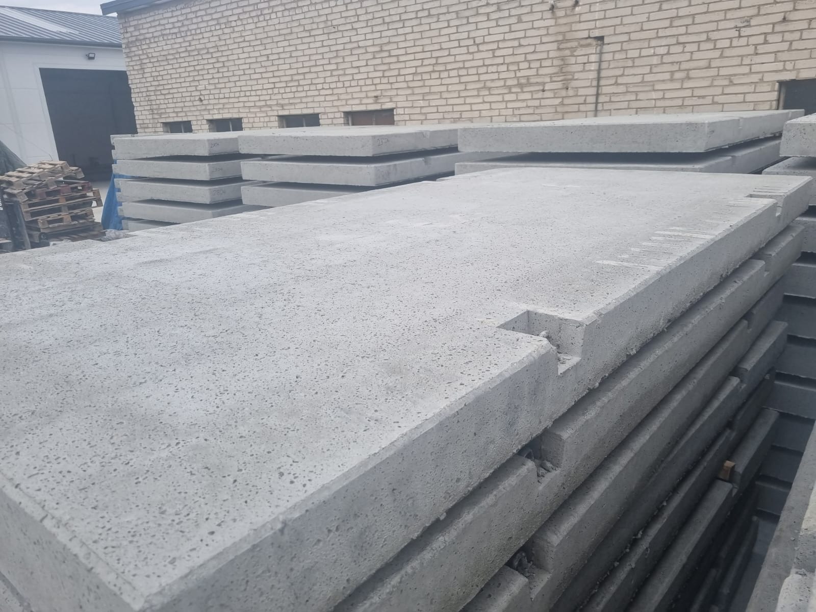 Stos szarych płyt drogowych z wycięciami na krawędziach, ułożonych na zewnątrz, z budynkiem z cegły w tle.