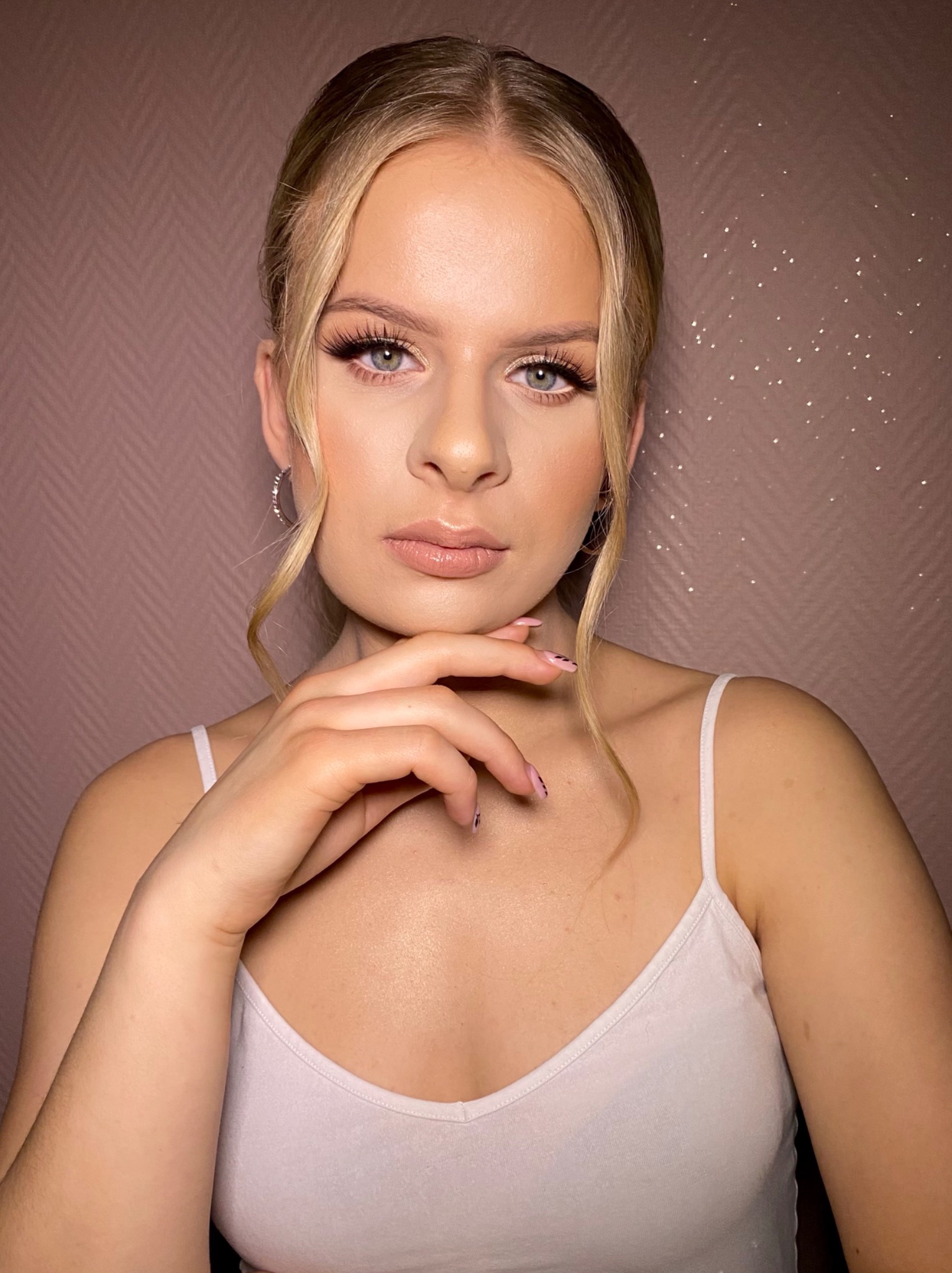 Portret młodej kobiety z delikatnym, naturalnym podkreśleniem urody, z uwzględnieniem przedłużonych rzęs i subtelnego manicure, na tle ściany z delikatnym wzorem.