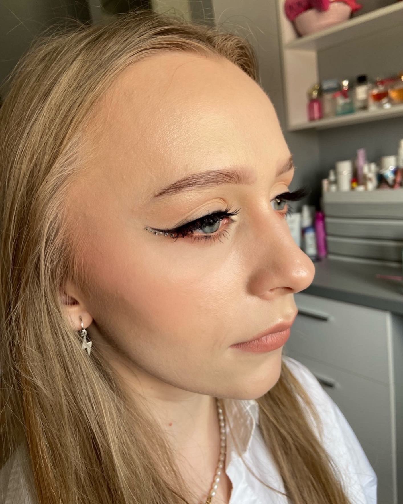 Profil młodej blondynki z delikatnym konturowaniem i czarnym eyelinerem ozdobionym drobnymi kryształkami, srebrny kolczyk w kształcie błyskawicy.