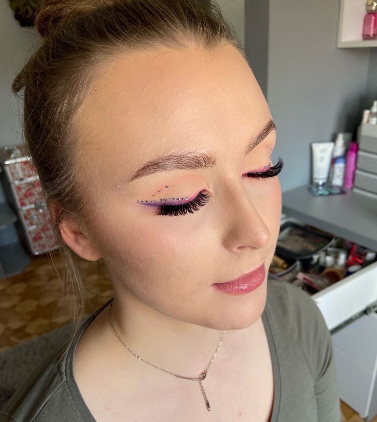 Portret kobiety z zamkniętymi oczami, prezentującej wyrazisty, artystyczny wizaż z fioletową kreską, sztucznymi rzęsami i drobnymi kryształkami wokół oczu.