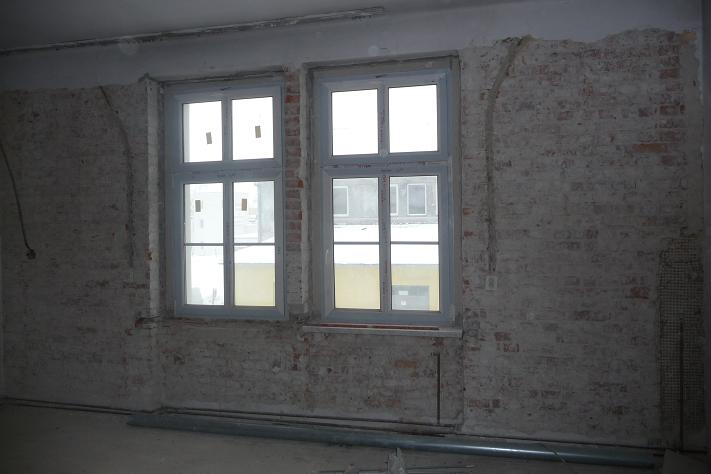 Wnętrze w trakcie remontu: surowe, otynkowane ściany z odsłoniętą cegłą, dwa okna z białymi ramami, widoczne przewody i rury na podłodze.
