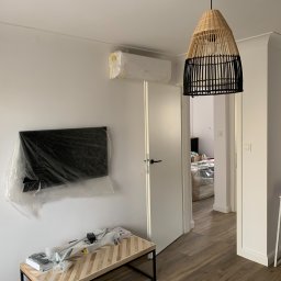 Milke Wykończenia - Białe wnętrze pokoju w trakcie remontu: klimatyzator i telewizor owinięte folią ochronną, drewniany stolik z akcesoriami, otwarta biała stolarka drzwiowa, wisząca lampa z abażurem z rattanu.