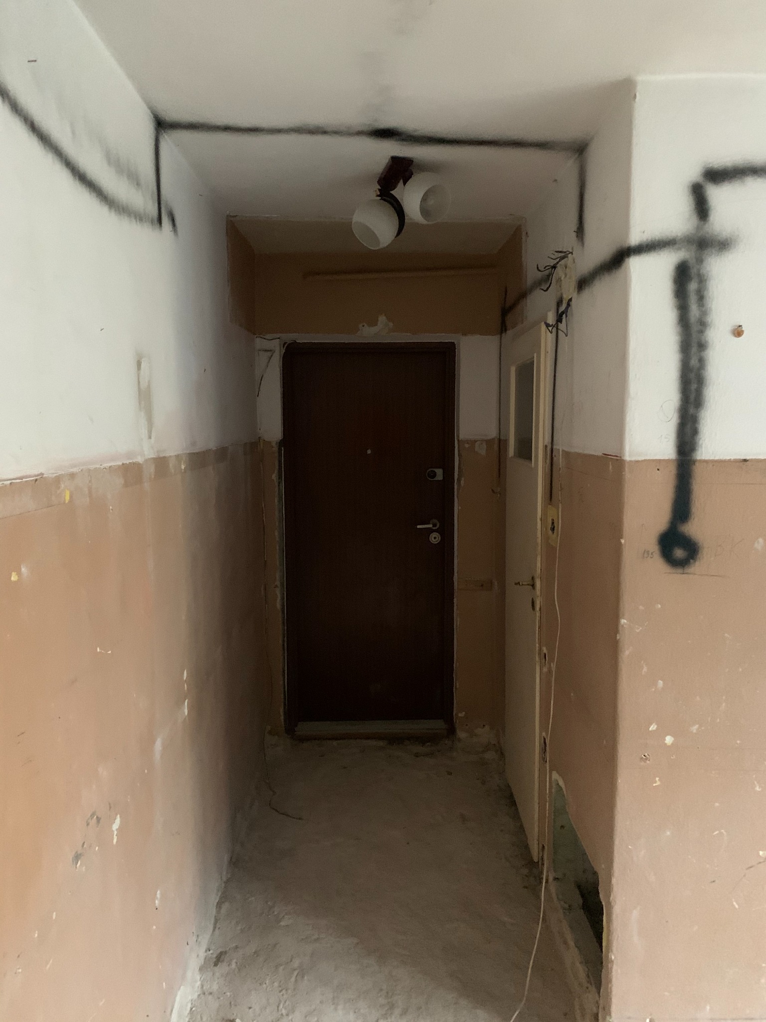 Korytarz w trakcie przygotowania do prac wykończeniowych: ściany częściowo pomalowane na beżowo, z widocznymi śladami po usuniętej farbie i odręcznymi czarnymi liniami na górnej części ścian...