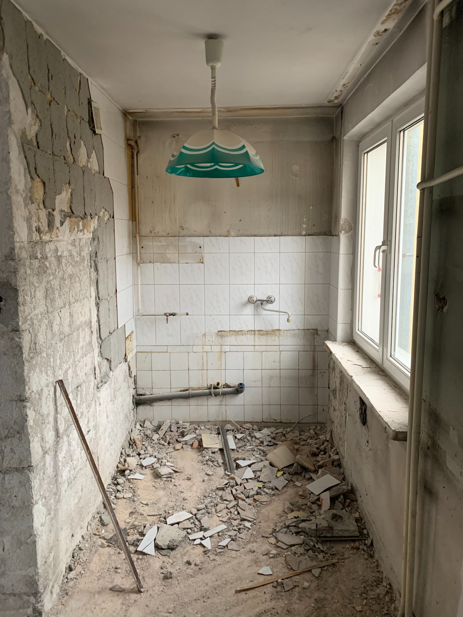 Łazienka w trakcie demontażu płytek i przygotowania do remontu, widoczne fragmenty starej instalacji, gruz na podłodze i lampa w stylu retro z zielonym abażurem.