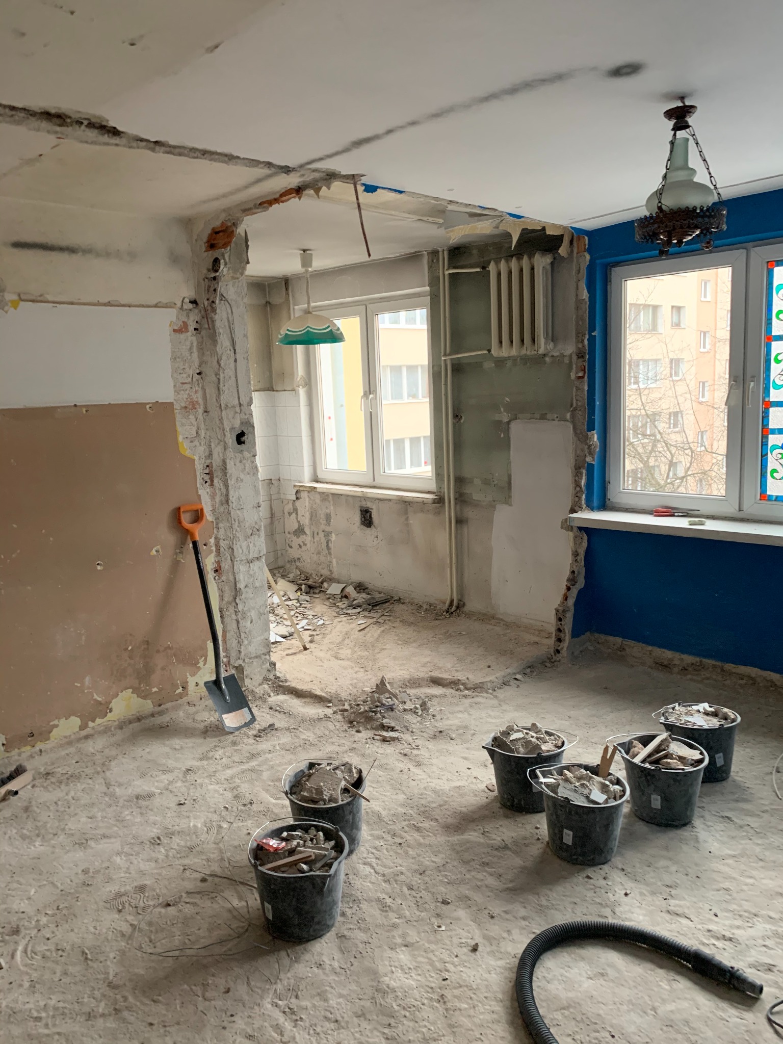 Wnętrze w trakcie demontażu: widoczne odsłonięte cegły, resztki tynku na ścianach, wiadra z gruzem na podłodze i łopata oparta o ścianę