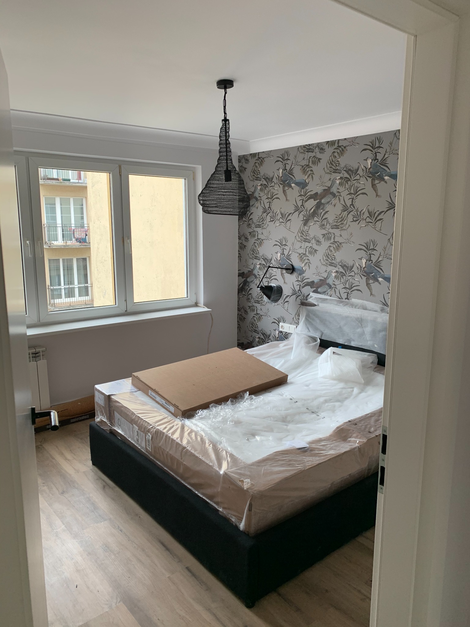 Sypialnia w trakcie urządzania: łóżko z czarną ramą, materac w folii, karton na materacu, wzorzysta tapeta z motywem ptaków i roślin, lampa wisząca z metalowej siatki, drewnopodobna podłoga.