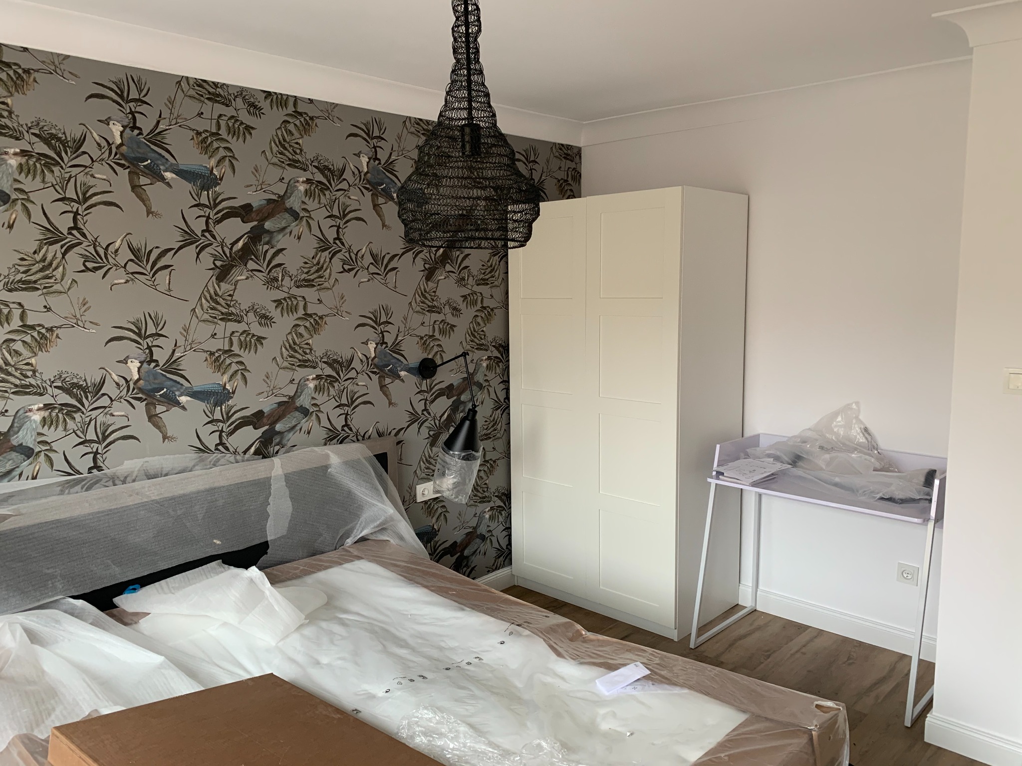 Pokój z tapetą w ptaki, zmontowaną szafą, biurkiem i łóżkiem w folii ochronnej, wisząca lampa z plecionki.