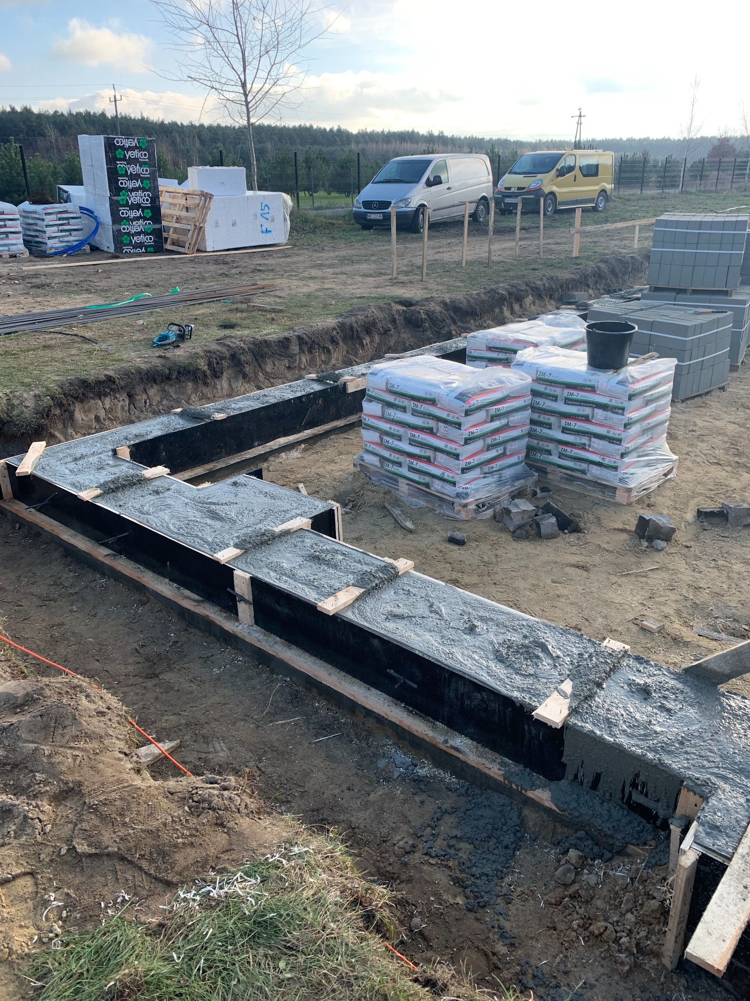 Wylany fundament pod budynek w kształcie litery L, widoczne szalunki z wypełnionym betonem, w tle palety z materiałami budowlanymi, worki z zaprawą murarską i dwa busy.