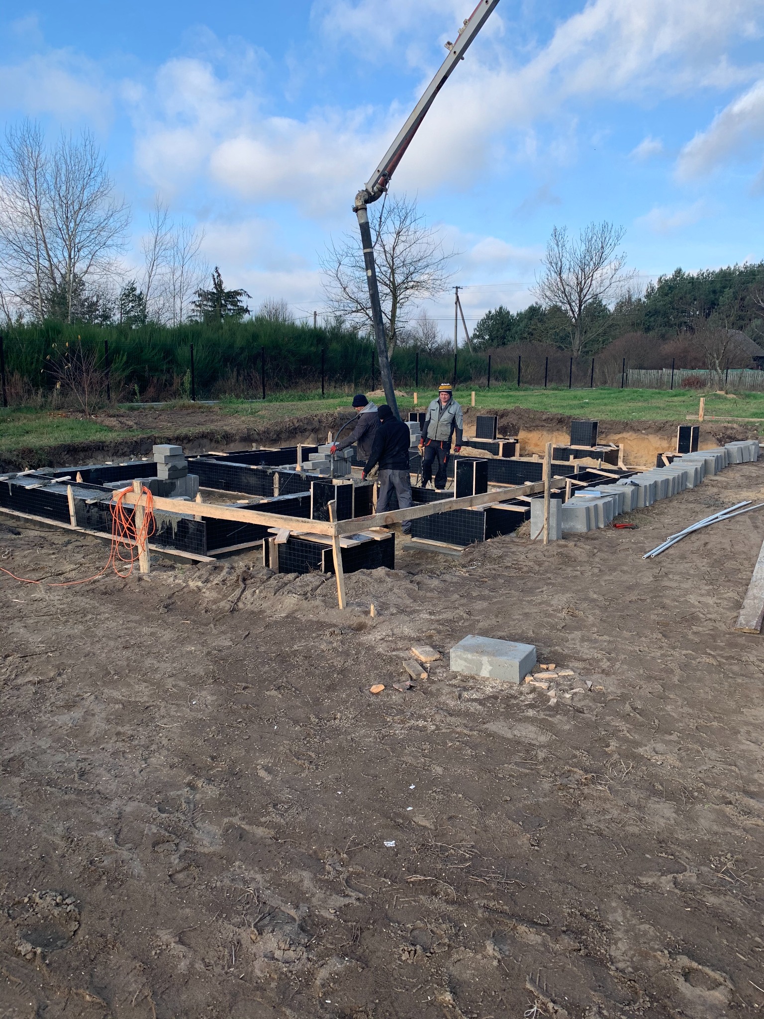 Wylewanie betonu do szalunków fundamentowych z użyciem pompy do betonu, widoczni dwaj pracownicy w odzieży roboczej, w tle drzewa i błękitne niebo.