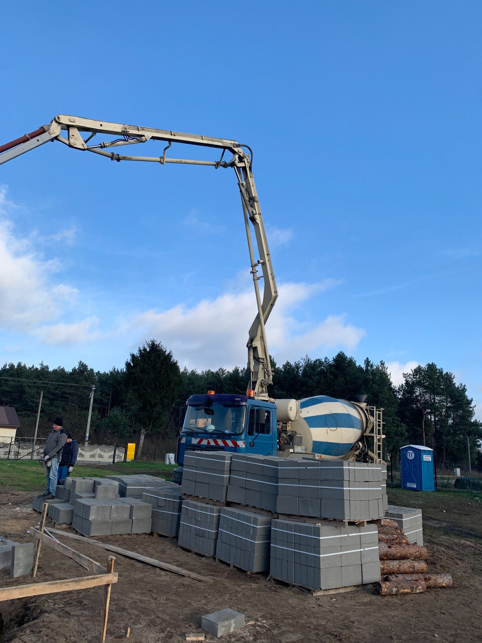 Na budowie, betonowa pompa z wysięgnikiem nad stertą szarych bloczków betonowych, w tle las i błękitne niebo z chmurami.