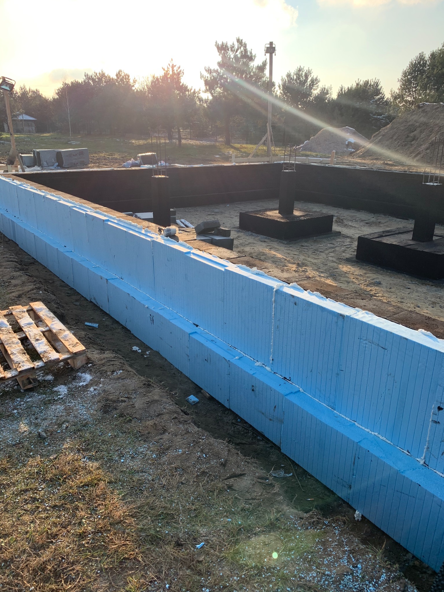 Fundamenty domu jednorodzinnego w trakcie budowy, ocieplone niebieskim styropianem, widoczne zbrojenia i drewniana paleta w pobliżu.