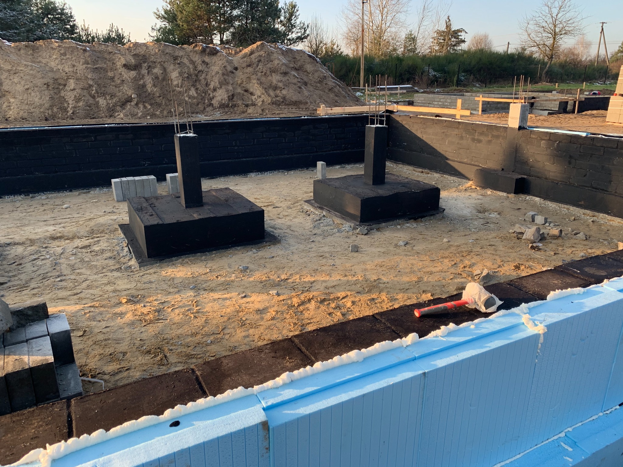 Fundamenty domu w trakcie budowy: zaizolowane ściany fundamentowe z wystającymi prętami zbrojeniowymi, podłoże piaszczyste, w tle pryzma ziemi.