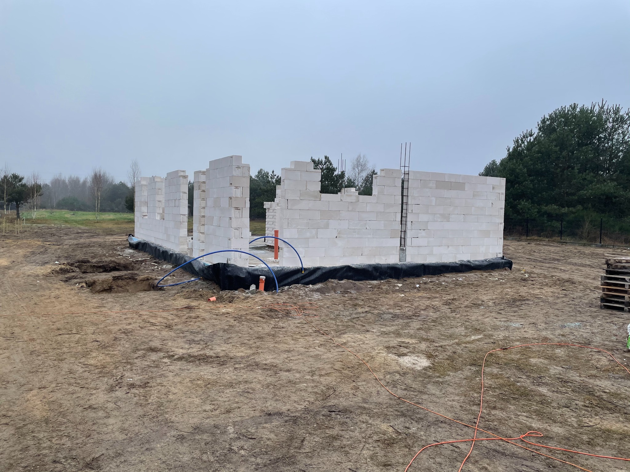 Wznoszenie ścian parteru domu jednorodzinnego z białych bloczków, widoczne zbrojenie, niebieskie rury instalacyjne i czarna folia hydroizolacyjna na fundamencie, teren budowy w pochmurny dzień.