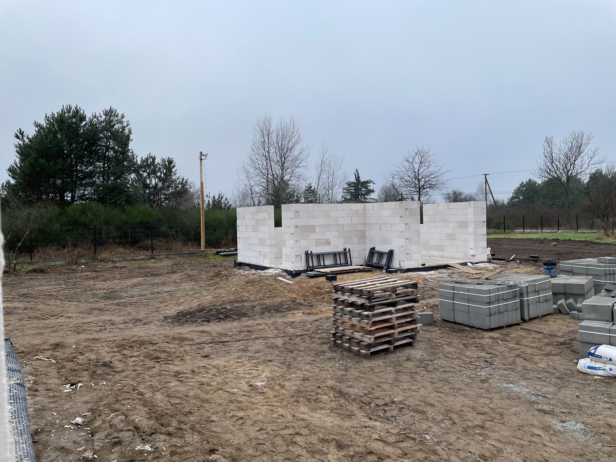 Wznoszone ściany domu z bloczków betonu komórkowego Ytong na działce z paletami i workami zaprawy, widok w pochmurny dzień.