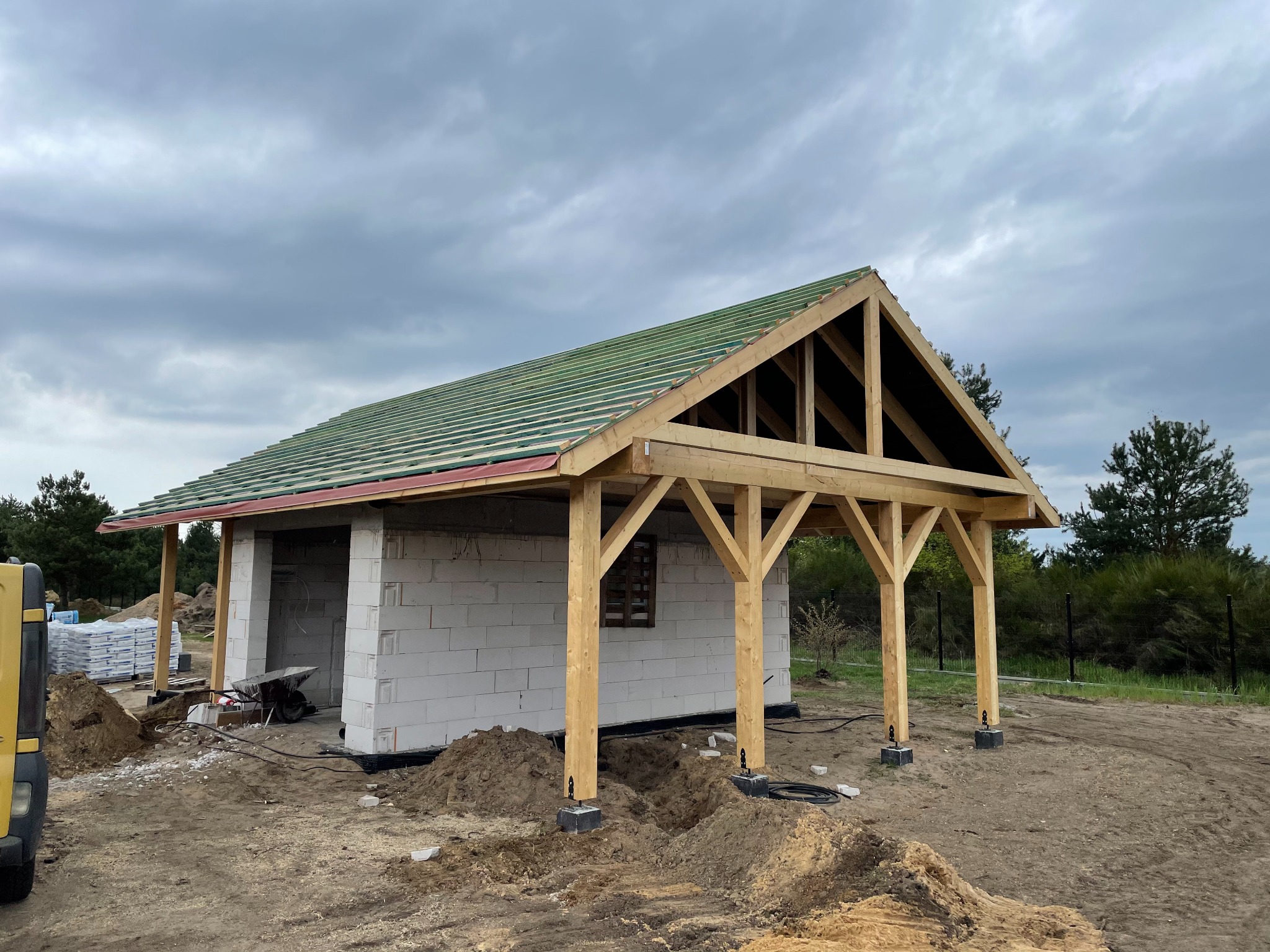 Budowa parterowego budynku z białych bloczków i drewnianą konstrukcją dachu pokrytą zieloną membraną, widoczne narzędzia budowlane i hałdy ziemi.