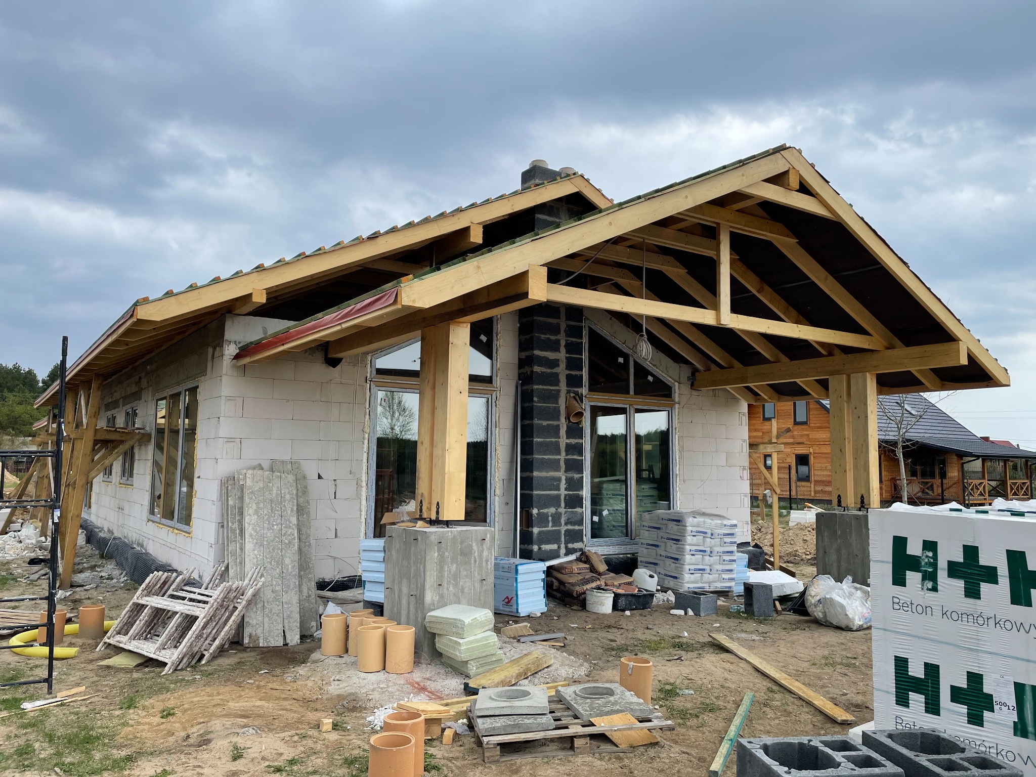 Budowa domu jednorodzinnego: stan surowy otwarty z widocznym drewnianym szkieletem dachu, bloczkami z betonu komórkowego i czarnym kominem z cegły klinkierowej na tle pochmurnego nieba.