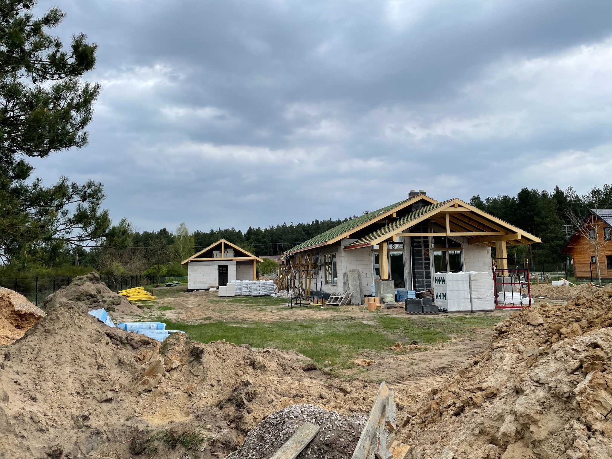 Budowa domu jednorodzinnego w stanie surowym otwartym, widoczne ściany z bloczków, konstrukcja dachu z drewna, stosy materiałów budowlanych i hałdy ziemi na działce.
