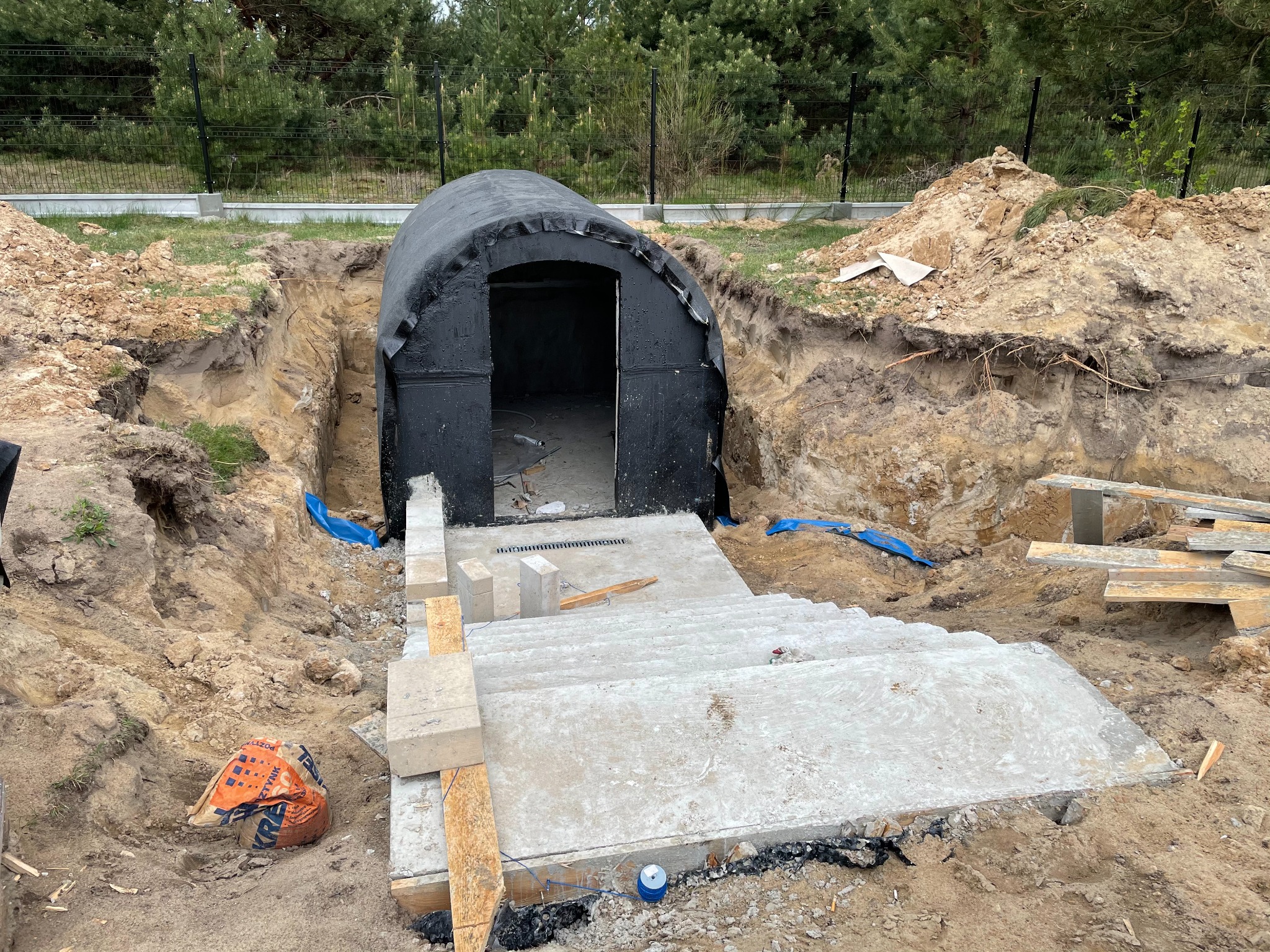 Budowa ziemianki: betonowe schody prowadzą do wejścia z czarną hydroizolacją, widoczny wykop w ziemi i stosy piasku, w tle zielone drzewa.