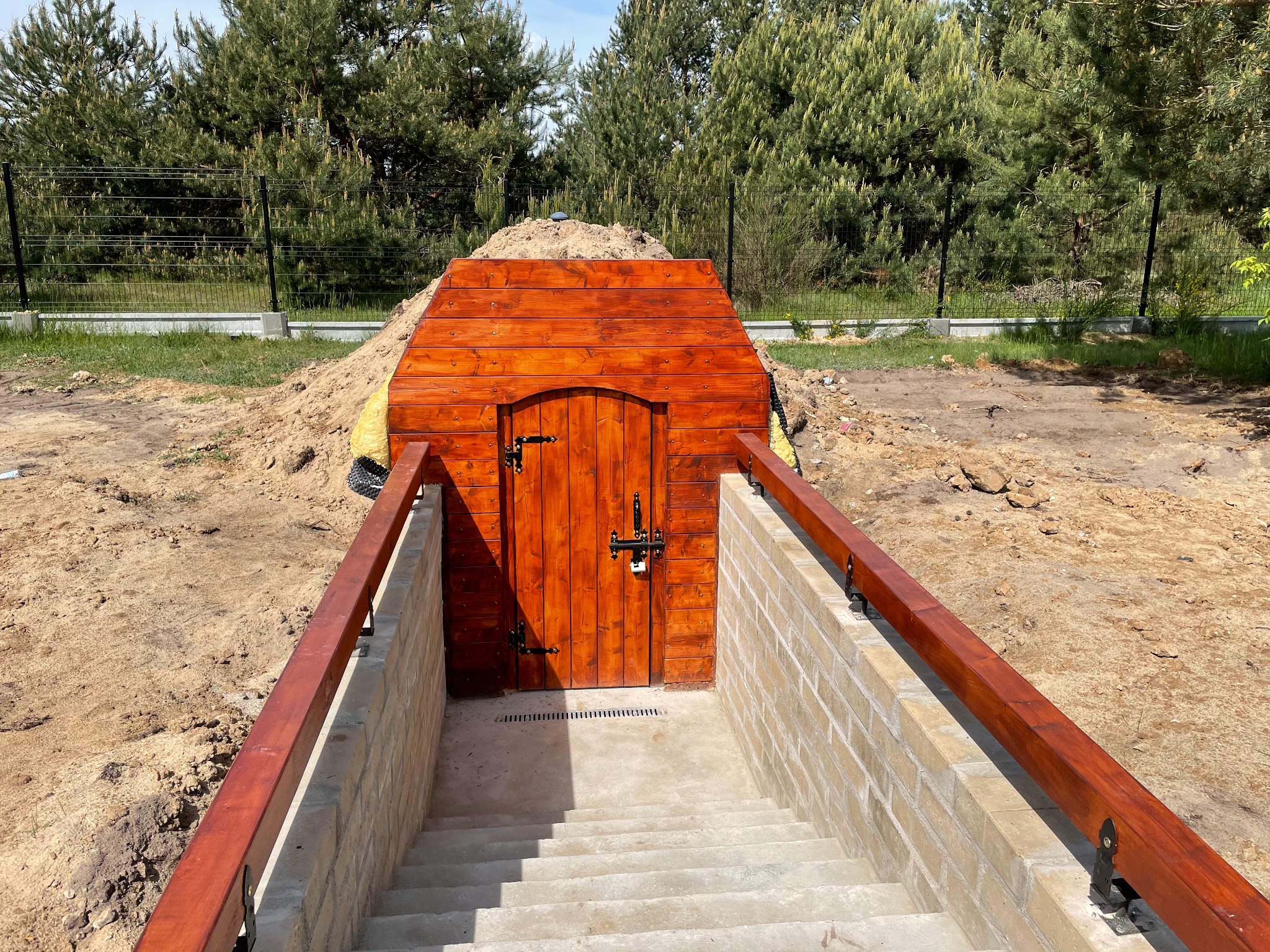 Wejście do ziemianki z drewnianymi drzwiami i schodami wyłożonymi jasną kostką brukową, otoczone ziemią i zielenią