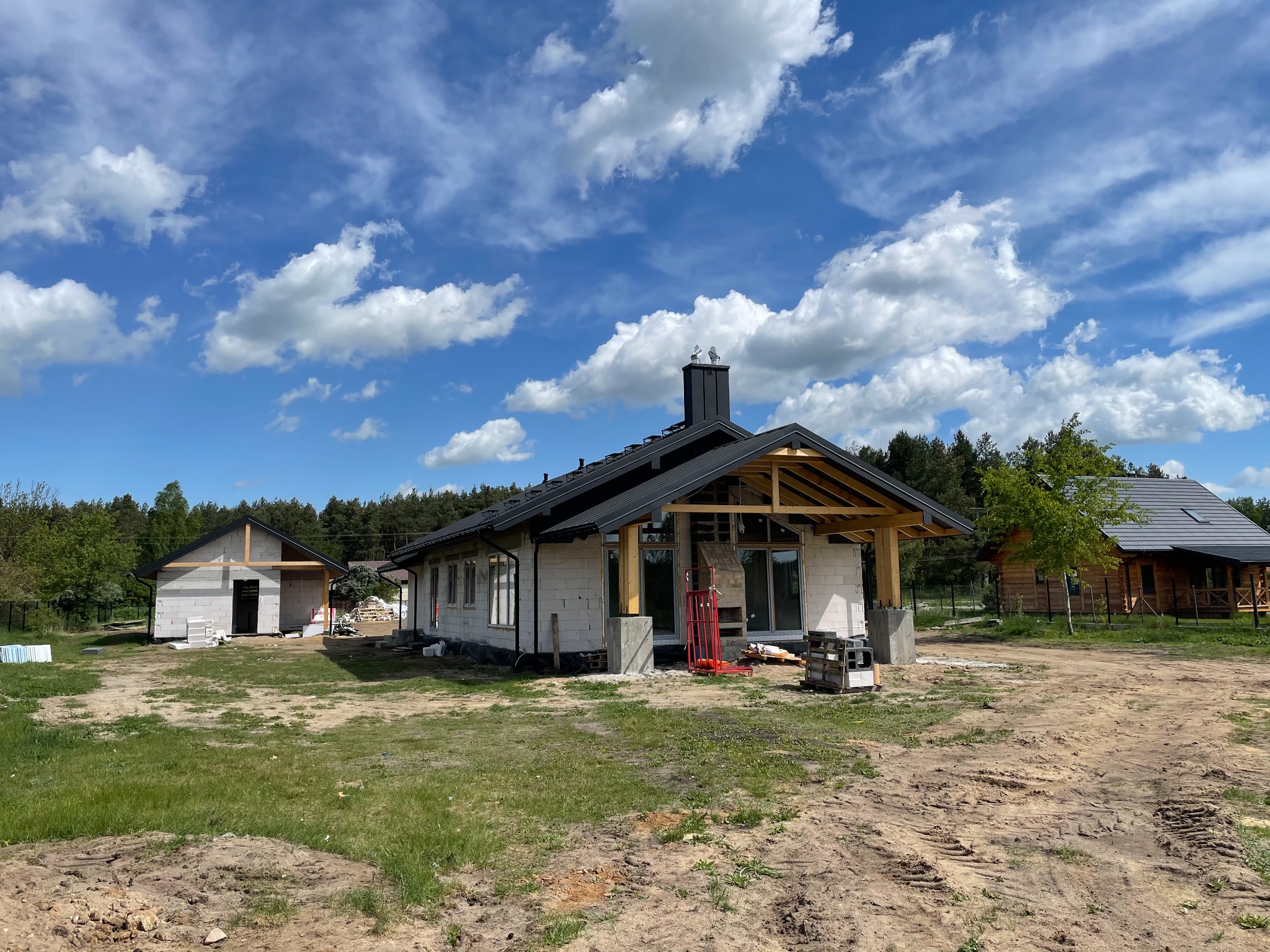 Budowa domu jednorodzinnego w stanie surowym otwartym, widoczne ściany z betonu komórkowego, drewniana konstrukcja dachu i komin, na działce drugi budynek w trakcie wznoszenia.