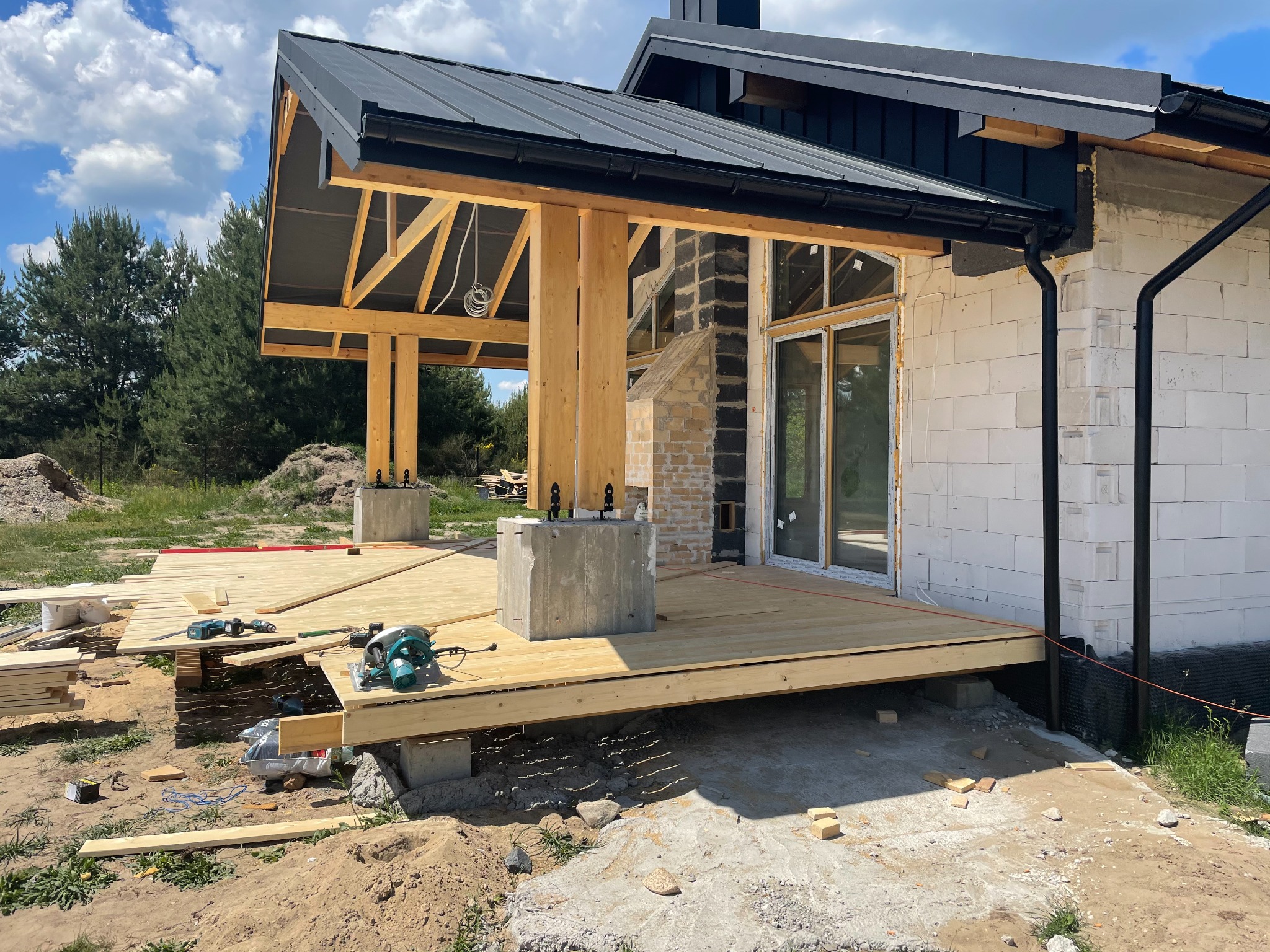 Budowa tarasu z drewnianą podłogą i zadaszeniem, widoczne narzędzia i materiały budowlane, ściana budynku z oknem w tle, etap prac wykończeniowych.