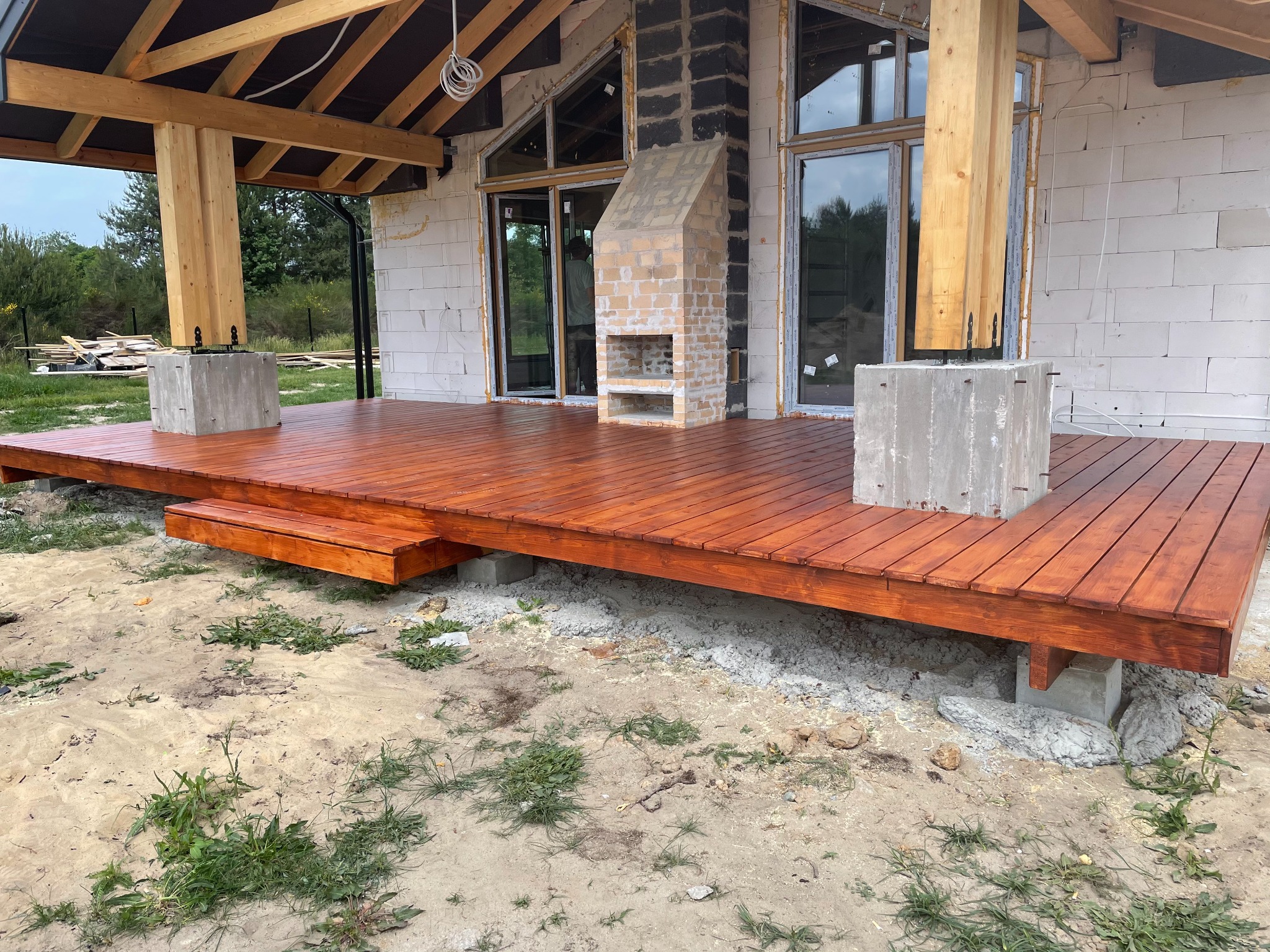 Nowo wybudowany, drewniany taras z brązową bejcą przy domu w stanie surowym, z widocznym kominkiem zewnętrznym z cegły i betonowymi podstawami słupów.