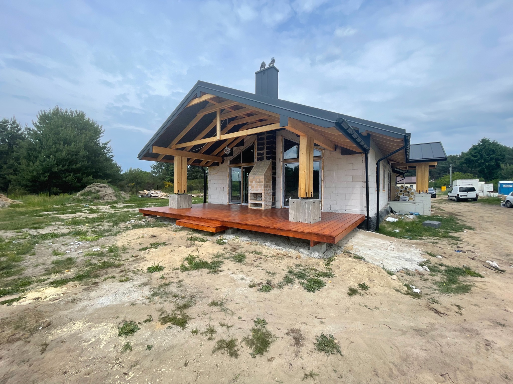 Dom jednorodzinny w trakcie budowy, widoczna konstrukcja dachu z drewnianymi belkami, białe ściany z pustaków, świeżo polakierowany drewniany taras i komin z ozdobnymi rzeźbami na szczycie.