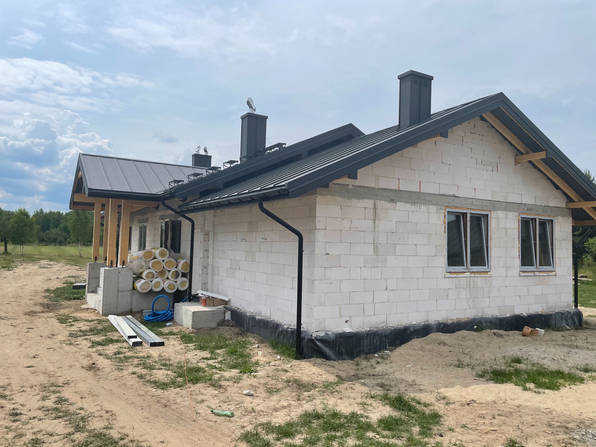 Budowa domu jednorodzinnego z białych bloczków, widoczna czarna blachodachówka, kominy, okna bez parapetów i ocieplenie w rolkach obok wejścia.