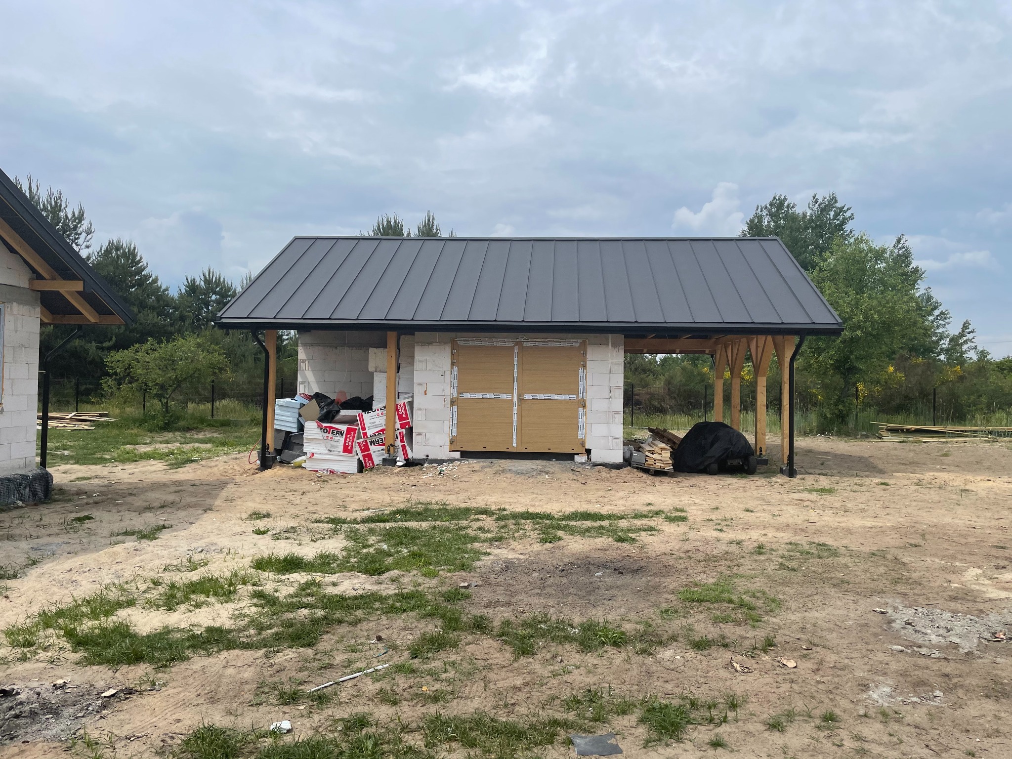 Budynek gospodarczy w trakcie budowy: ściany z bloczków, grafitowy dach z blachy na rąbek stojący, drewniane słupy podtrzymujące zadaszenie, styropian elewacyjny w paczkach obok.