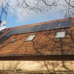 Czerwony dach z dachówki ceramicznej z zamontowanymi panelami słonecznymi i oknami dachowymi, widoczny komin z anteną satelitarną na tle błękitnego nieba z chmurami.