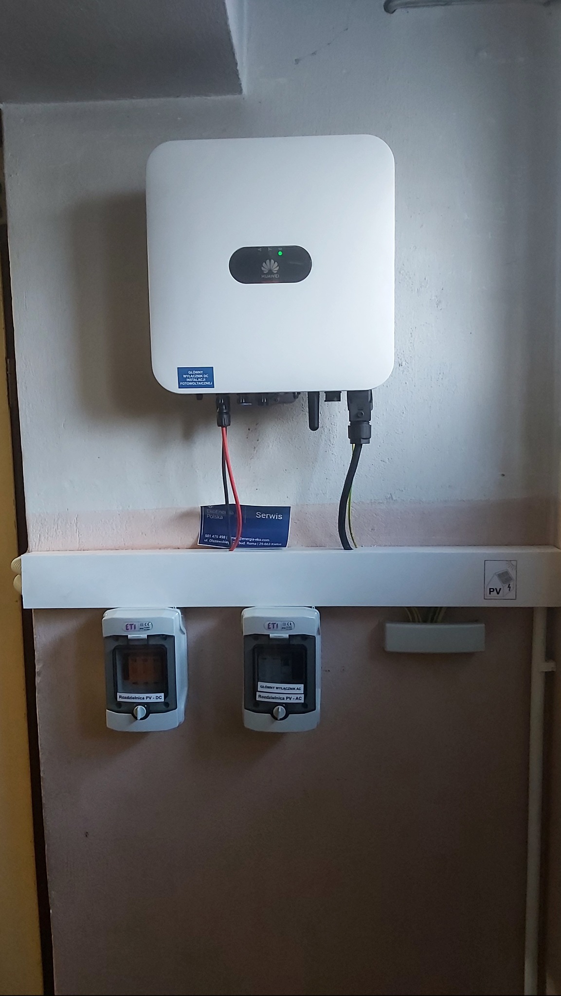 Biały inwerter Huawei zamontowany na ścianie, z widocznymi kablami, bezpiecznikami DC i AC w szarych obudowach ETI, oraz naklejką informacyjną o wyłączniku DC, zainstalowany wewnątrz budynku.