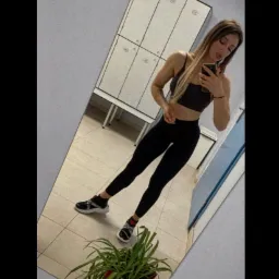 Kobieta w sportowym stroju robi selfie w lustrze siłowni, widoczne szafki, płytki podłogowe i roślina doniczkowa w rogu.