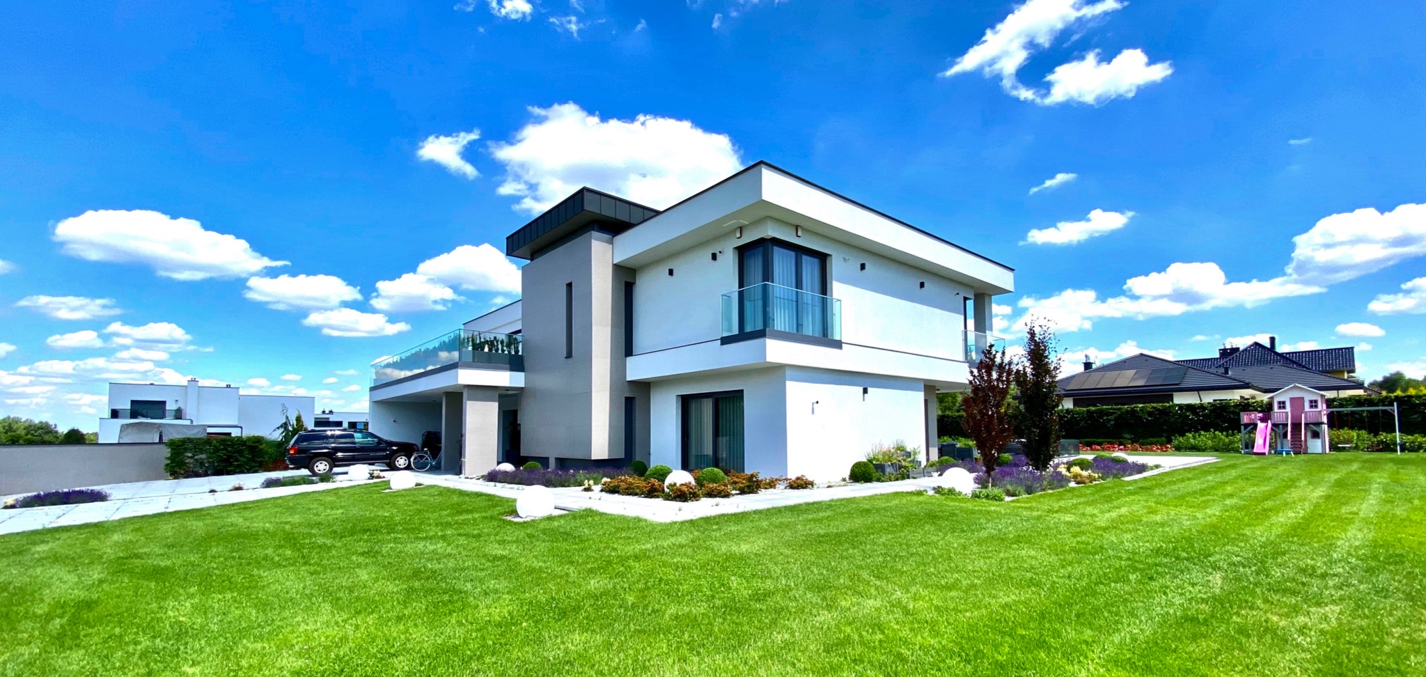 Nowoczesny dom jednorodzinny z ogrodem, biała elewacja, szklane balustrady balkonowe, równo przystrzyżony trawnik, białe lampy kuliste, samochód w garażu, błękitne niebo z chmurami.