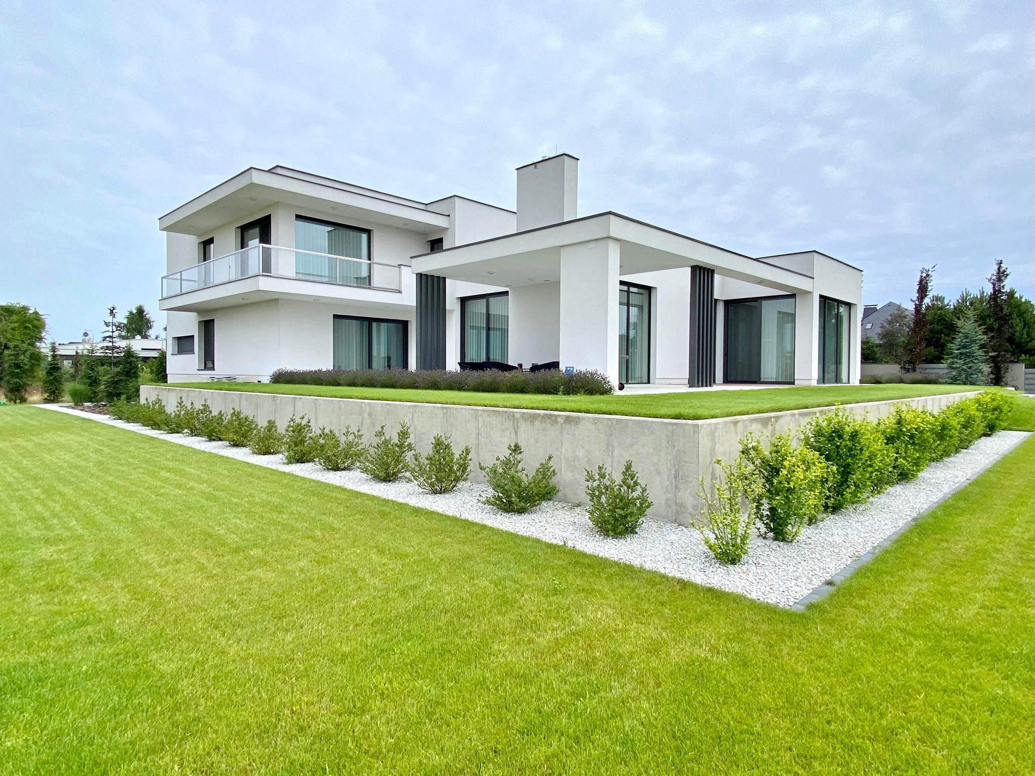 Nowoczesny, biały dom jednorodzinny z płaskim dachem i dużymi oknami, otoczony zadbanym trawnikiem z ozdobnymi krzewami oddzielonymi białym żwirem.