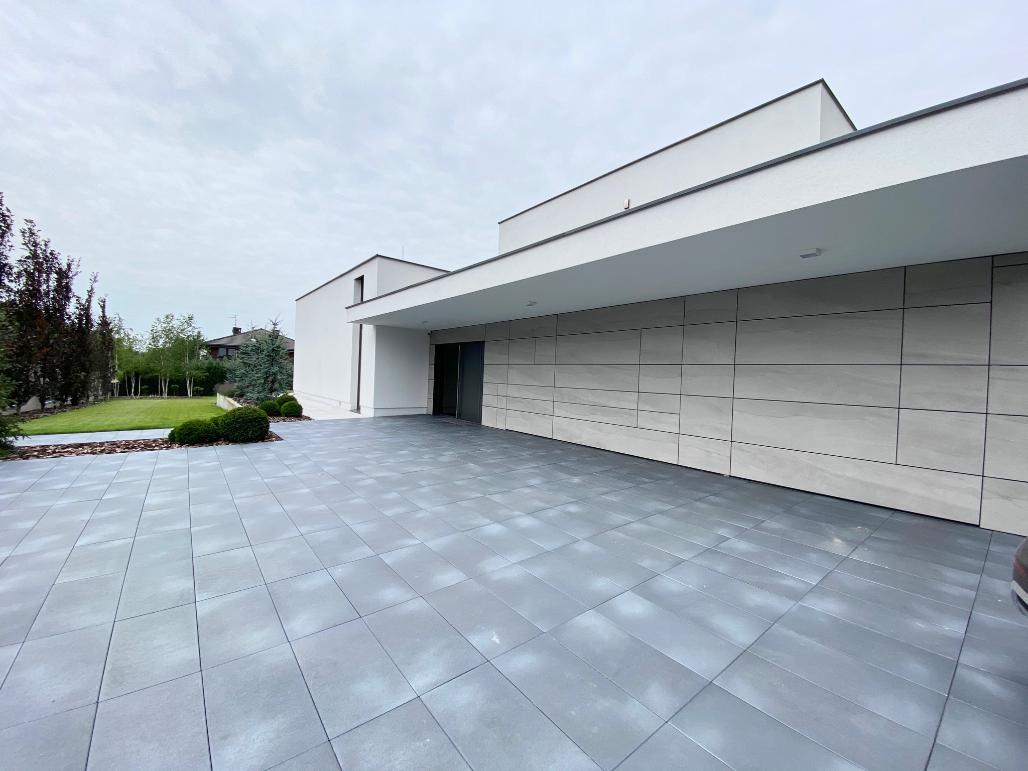 Nowoczesny dom jednorodzinny z szarą elewacją i wyłożonymi płytami chodnikowymi, widok z zewnątrz w pochmurny dzień.