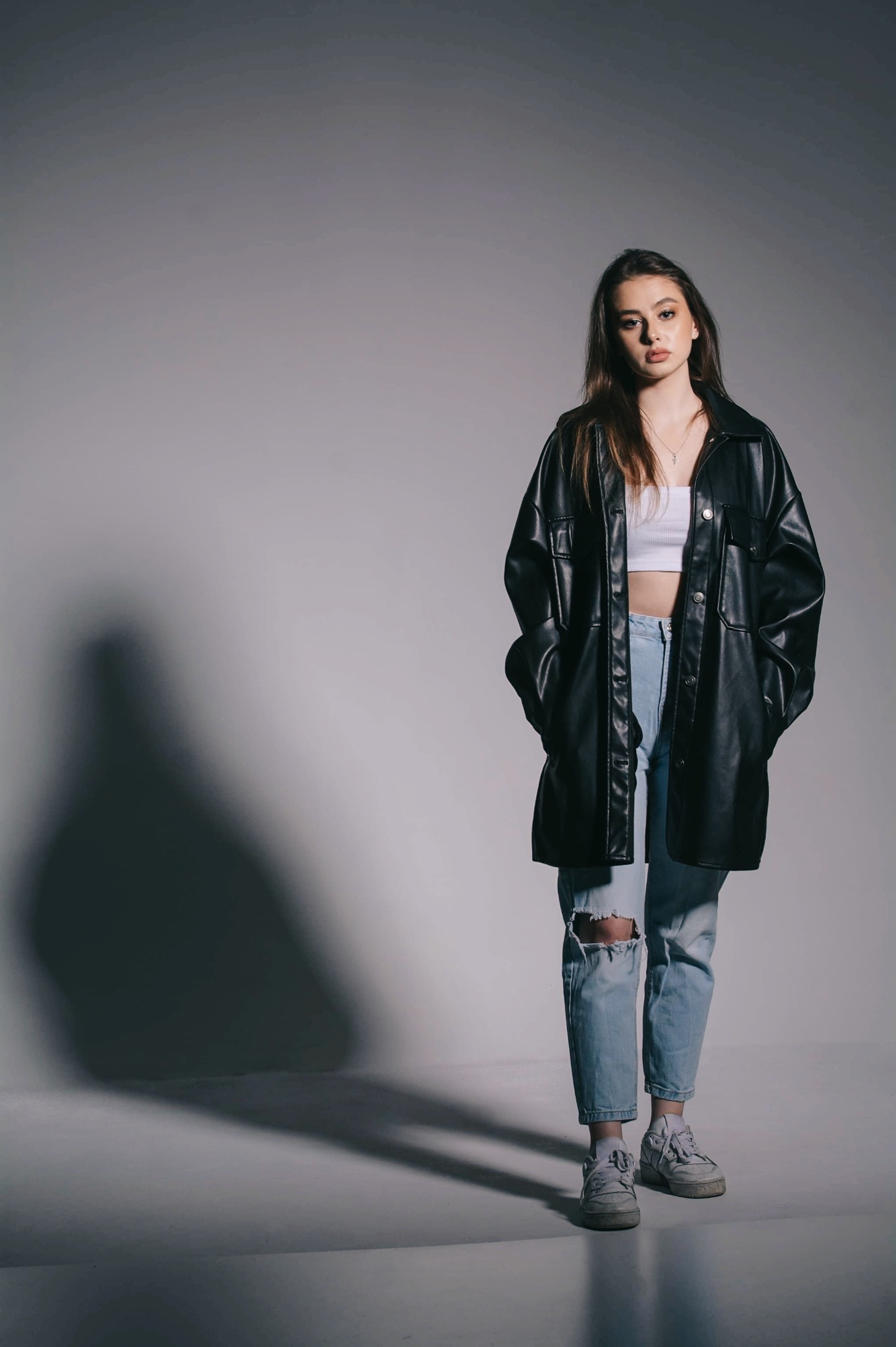Młoda kobieta w stylizacji z czarną, skórzaną kurtką, białym topem i podartymi jeansami, pozująca na szarym tle z wyraźnym cieniem