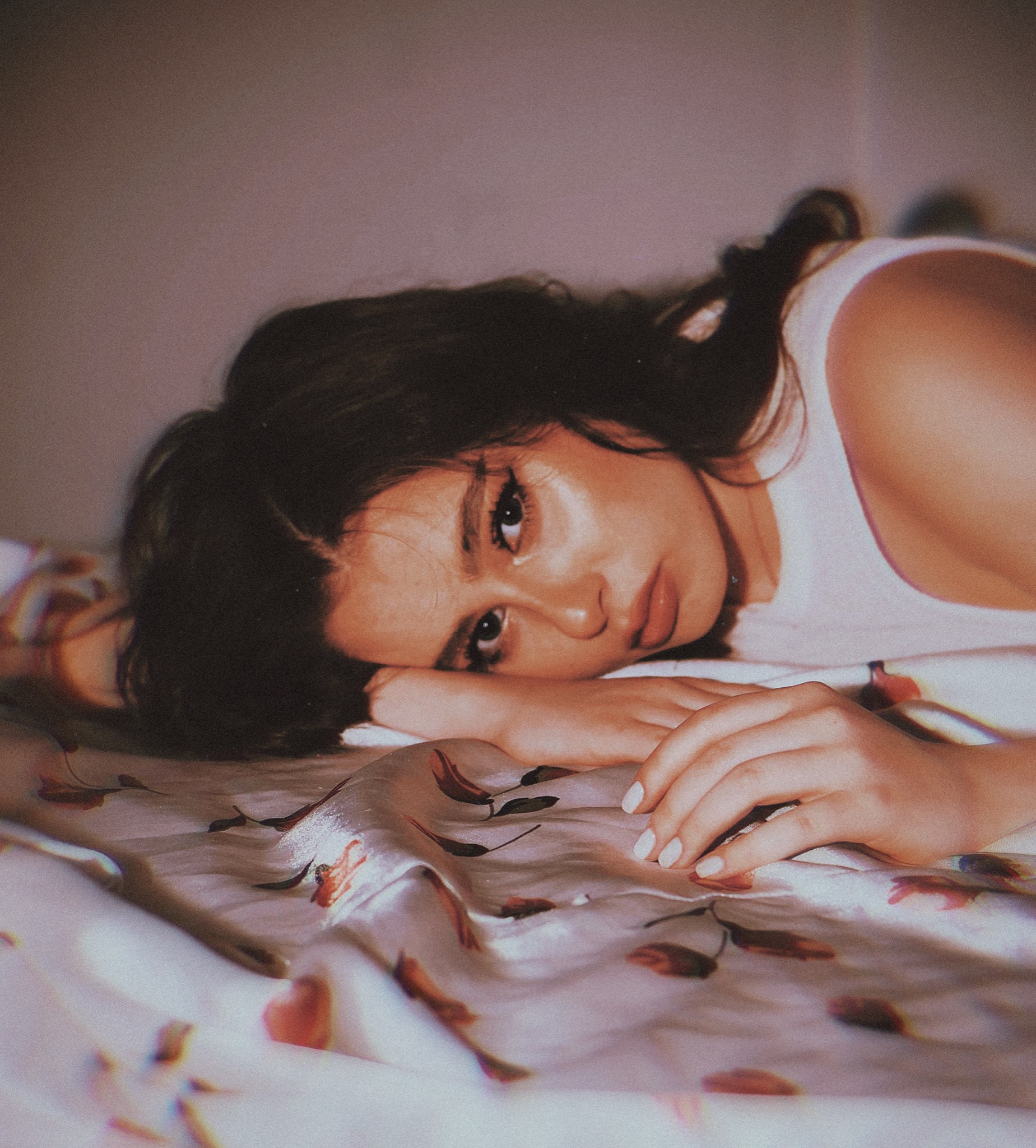 Portret kobiety leżącej na wzorzystej, satynowej pościeli, z ręką pod policzkiem, w naturalnym makijażu i białej koszulce na ramiączkach.