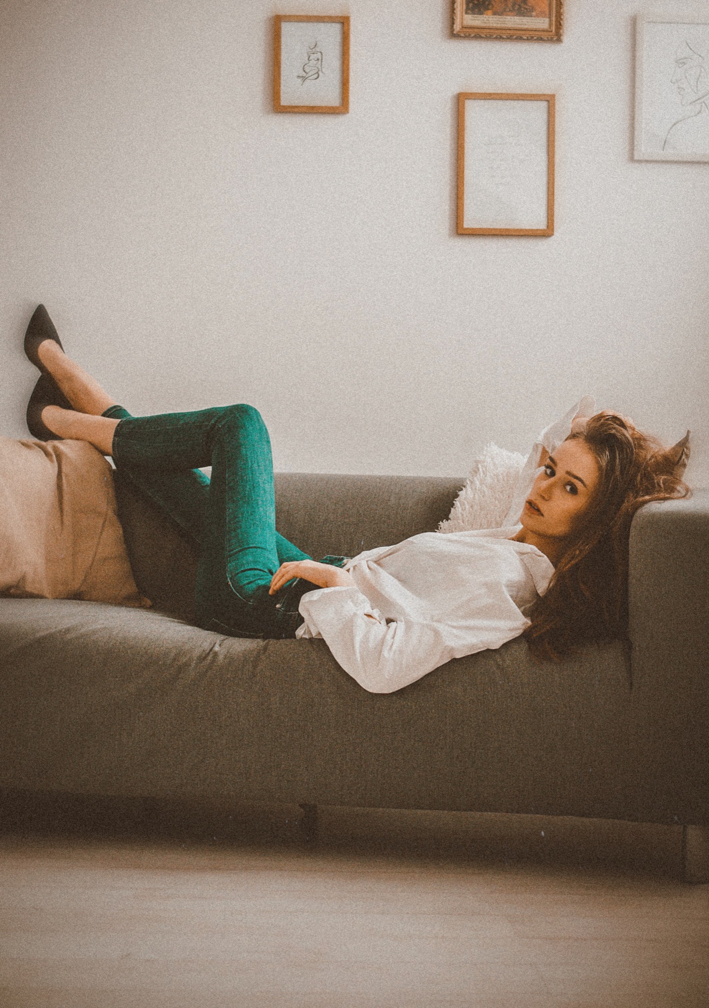 Portret kobiety w białej koszuli i zielonych dżinsach leżącej na szarej sofie na tle białej ściany z ramkami.