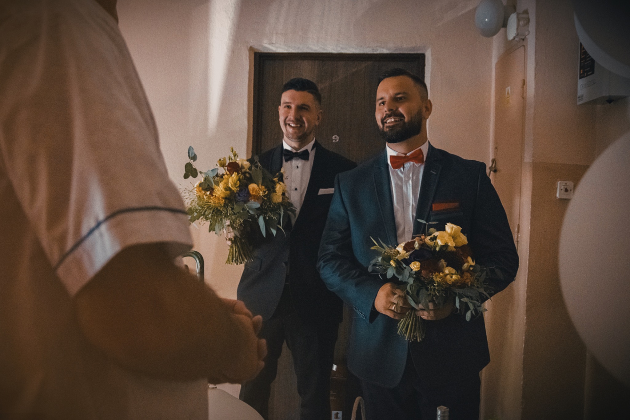 Uśmiechnięci panowie w garniturach z bukietami w dłoniach, jeden z czerwoną muchą, stojący przed drzwiami z numerem 9, widok zza ramienia osoby w piżamie.